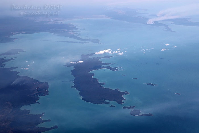 Flying out towards the Arafura Sea looking at Inglis Island, just off the coast of East Arnhem Land which is home of the Yolngu People, near Nhulunbuy in the Northern Territory. Traditional fire stick burning, an indigenous fire management practice is seen in the distance on the mainland and on the distant Elcho Island. 11°57'26.4"S 136°23'12.9"E. High altitude shots from a flight between Sydney Australia and Manila, Philippines.